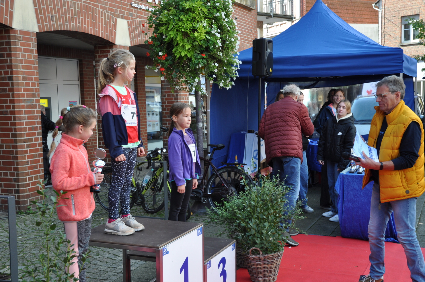 Drei Mädchen stehen auf dem Siegertreppchen bei einer Sportveranstaltung, während ein Mann ihnen eine Urkunde überreicht.