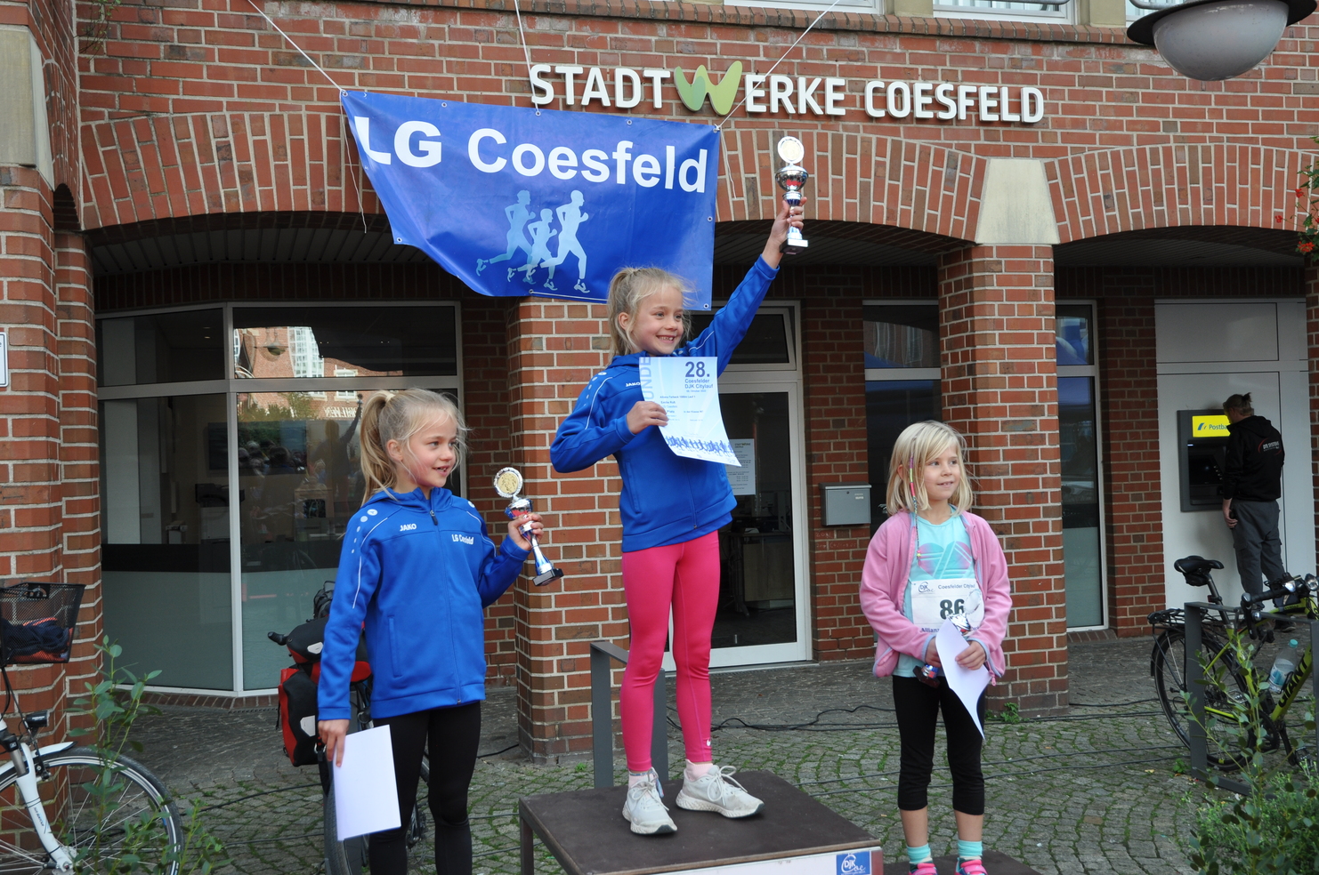 Drei Mädchen stehen auf dem Siegertreppchen, feiern ihre Platzierungen mit Pokalen und einer Urkunde vor einem Gebäude.