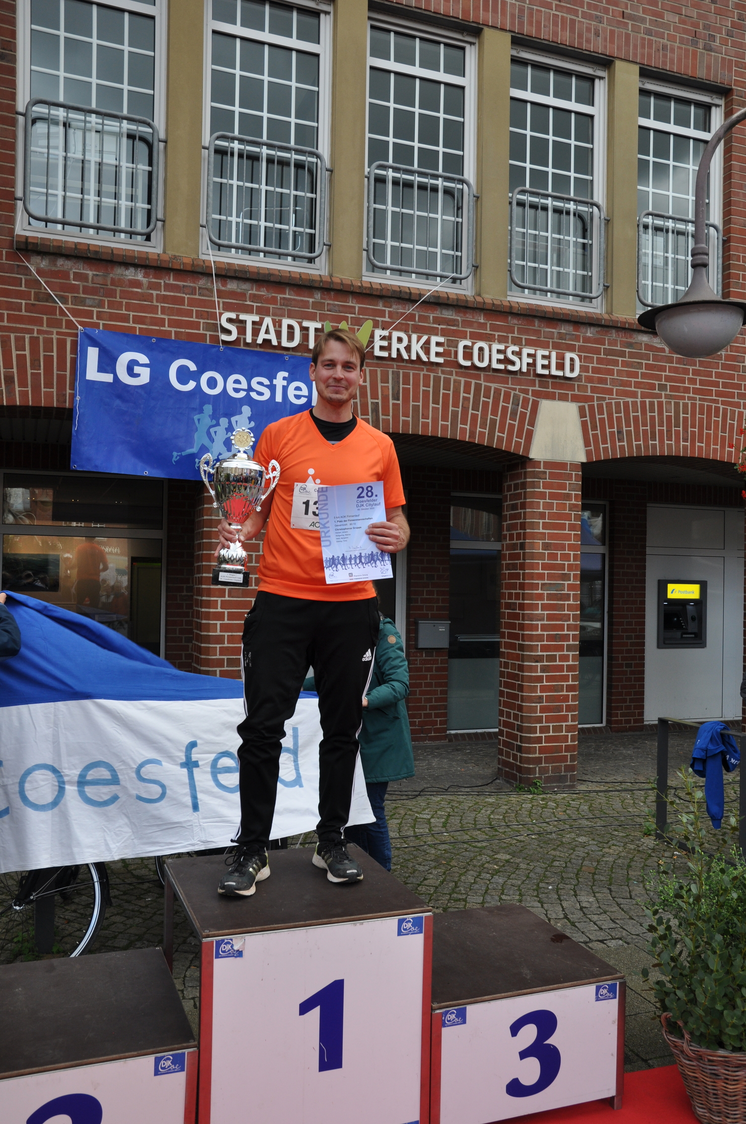 Mann in orangeem Shirt steht auf dem Siegerpodest mit Pokal und Urkunde vor der Gebäude-Fassade in Coesfeld.