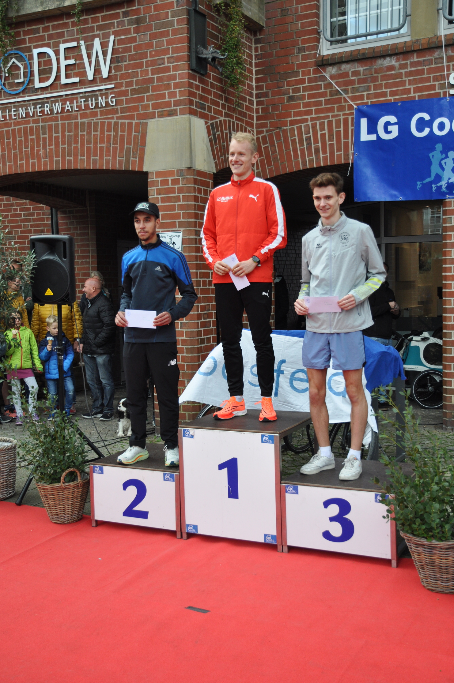 Drei Läufer stehen auf dem Siegertreppchen mit Platznummern, während Zuschauer im Hintergrund applaudieren.