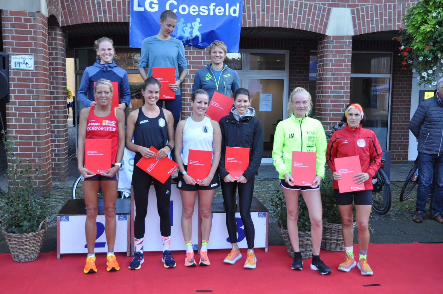 Gruppenfoto von Läuferinnen mit Urkunden bei der Siegerehrung vor einem Gebäude, roter Teppich und LG Coesfeld Banner im Hintergrund.