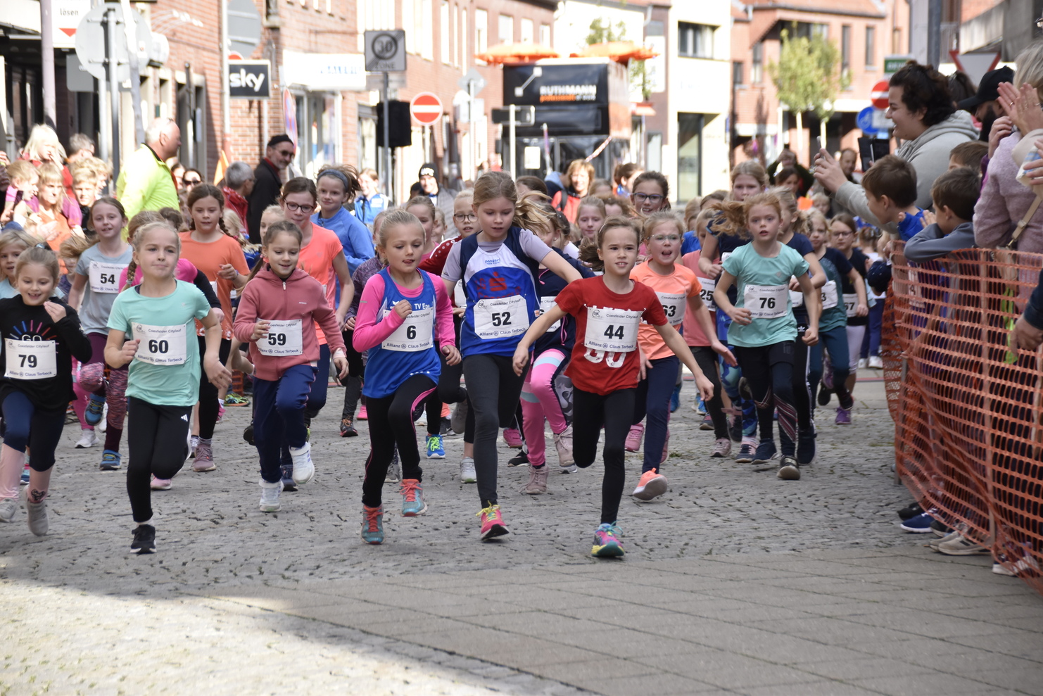Gruppe junger Mädchen in Sportbekleidung beim Start eines Rennen auf einer gepflasterten Straße, Zuschauer im Hintergrund.