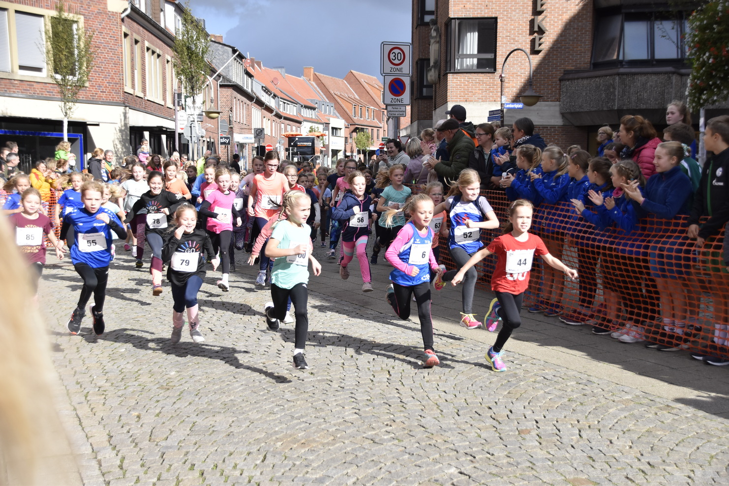 Gruppe von Mädchen in Sportkleidung bei einem Laufereignis auf einer gepflasterten Straße, umgeben von Zuschauern.