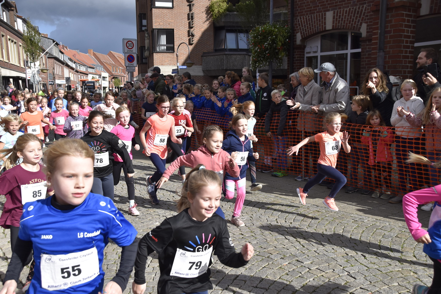 Eine Gruppe junger Läuferinnen startet beim Stadtlauf, umgeben von zuschauenden Menschen und einer festlichen Stimmung.