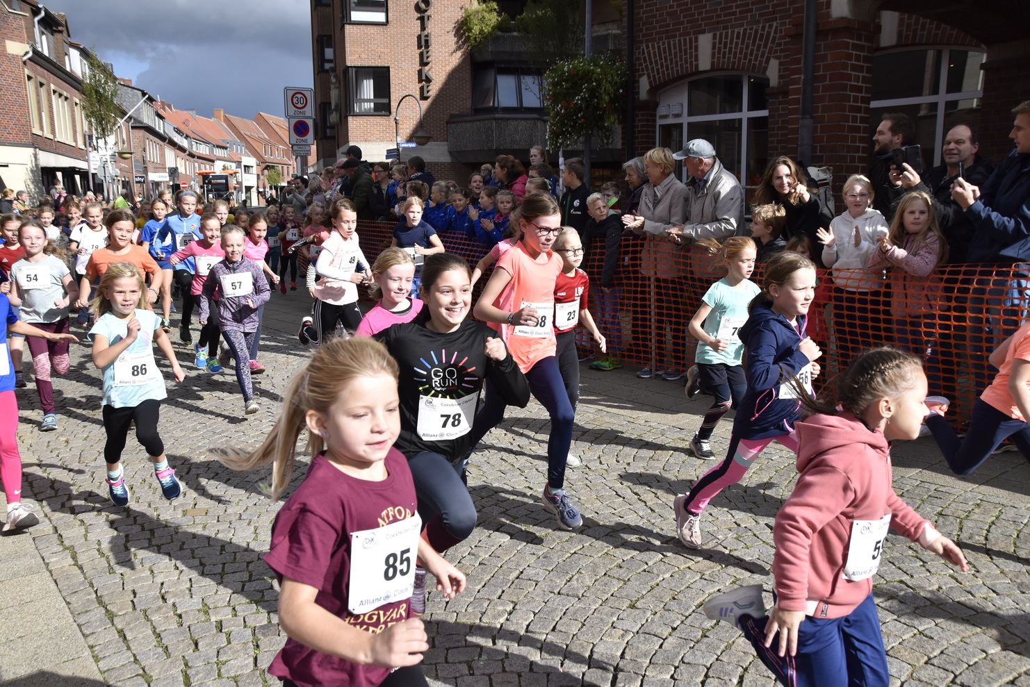 Eine Gruppe von Kindern läuft fröhlich einen Wettlauf auf einer befestigten Straße, Zuschauer jubeln entlang der Strecke.