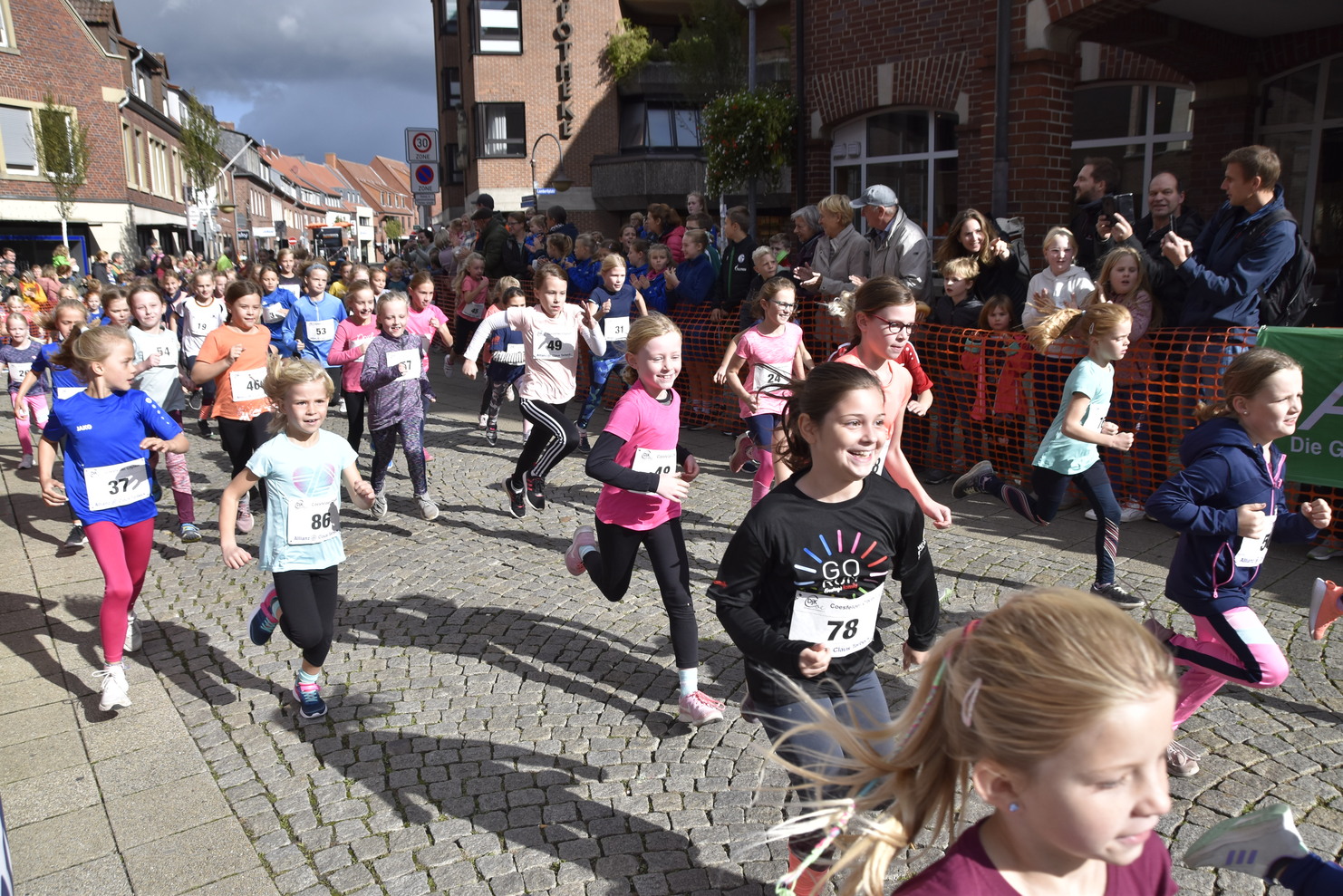 Mädchen in Sportkleidung laufen begeistert bei einem Kinderlauf durch eine belebte Straße mit Zuschauern.