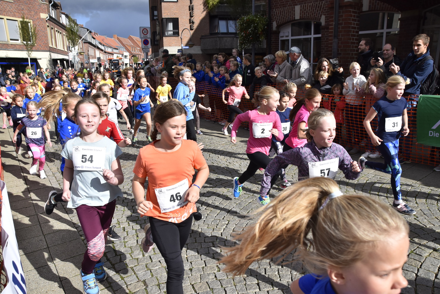 Gruppe von Mädchen läuft fröhlich einen Straßenlauf, umgeben von Zuschauern auf einer gepflasterten Straße.