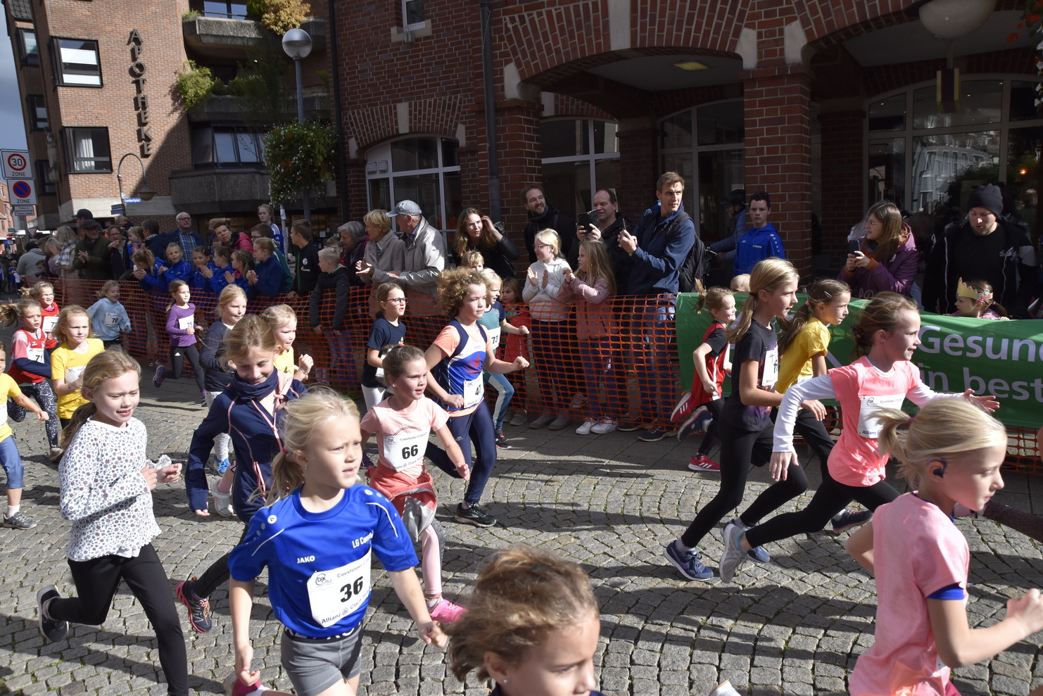 Gruppierung von Kindern beim Laufwettbewerb auf einer Straße, umgeben von Zuschauern und orangefarbener Absperrung.