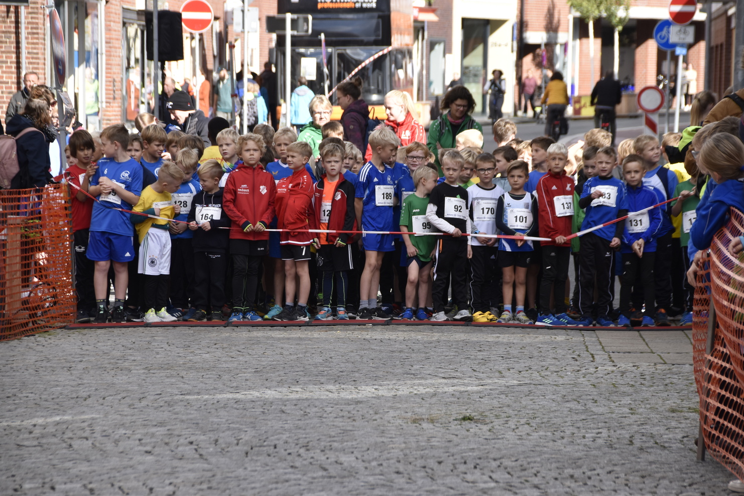Gruppierung von Kindern in Sportbekleidung mit Startnummern an der Startlinie eines Rennens auf einer gepflasterten Straße.