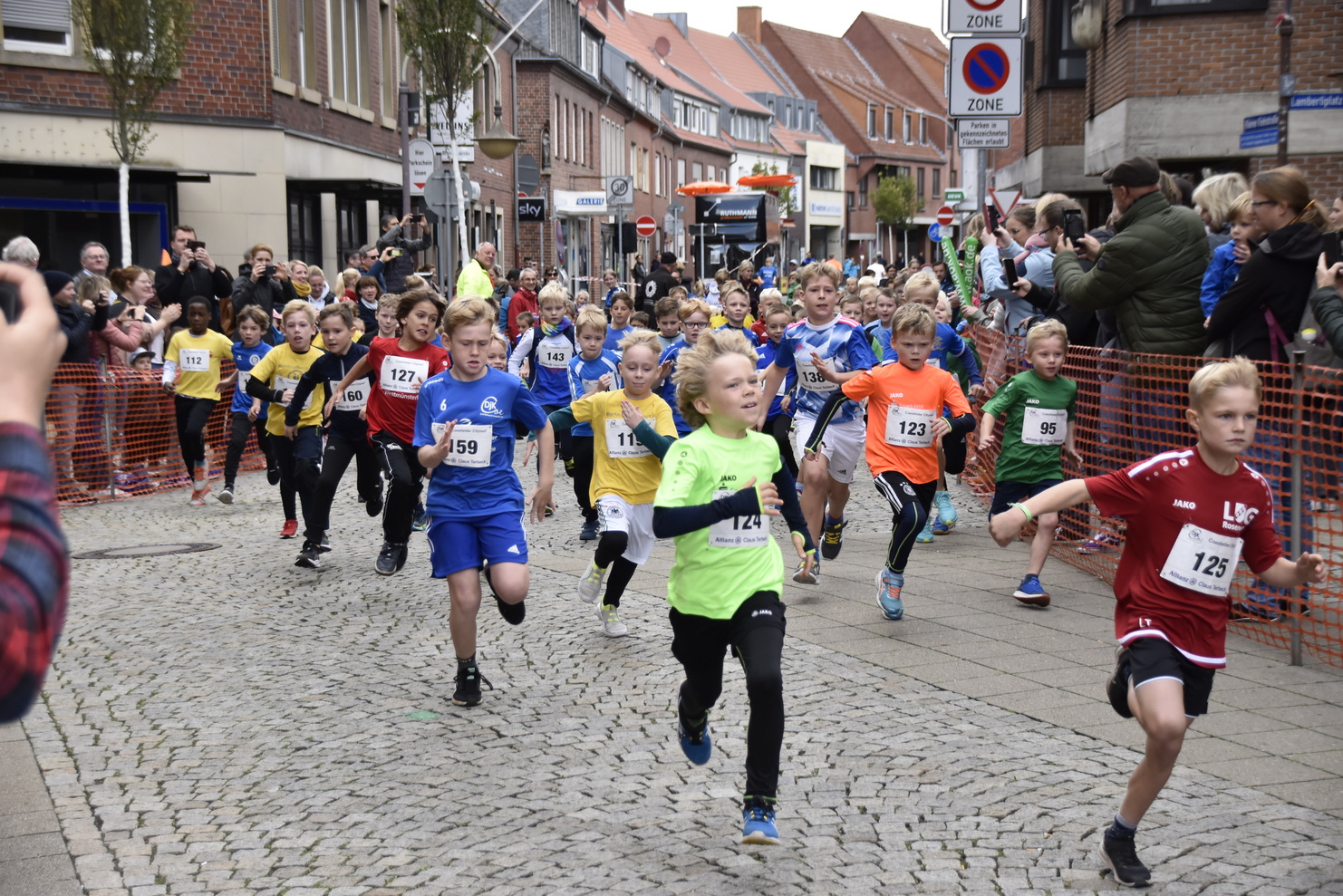 Gruppe von Kindern in bunten Lauftrikots bei einem Wettlauf auf einer gepflasterten Straße, Zuschauer beobachten.