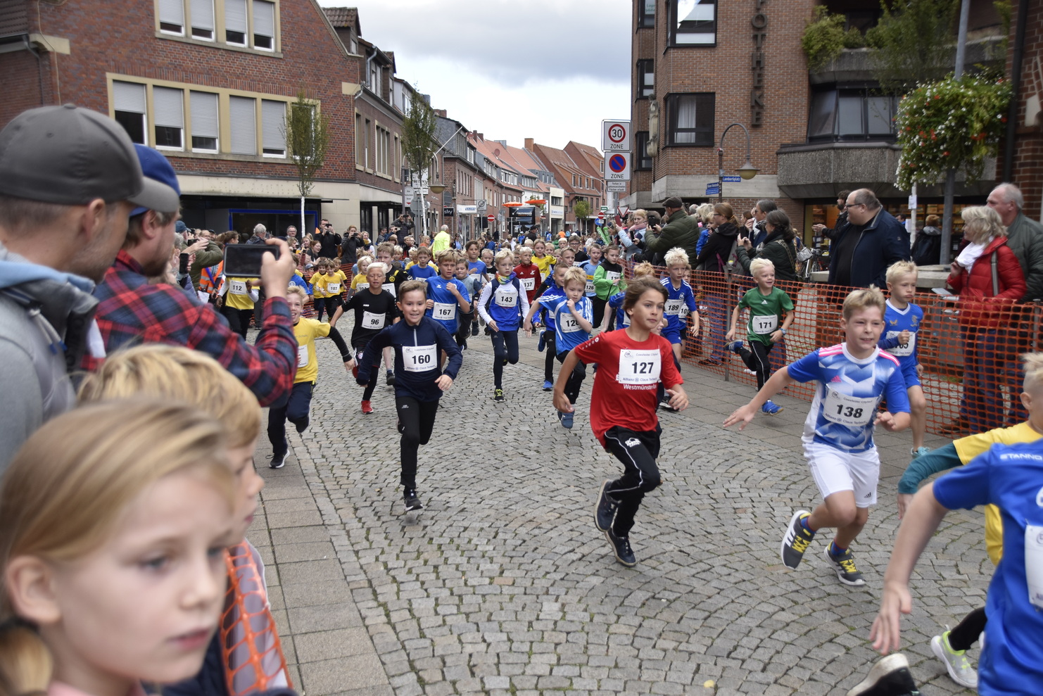 Kinder in Sportbekleidung laufen mit Freude eine Straße entlang, während Zuschauer entlang der Strecke stehen.