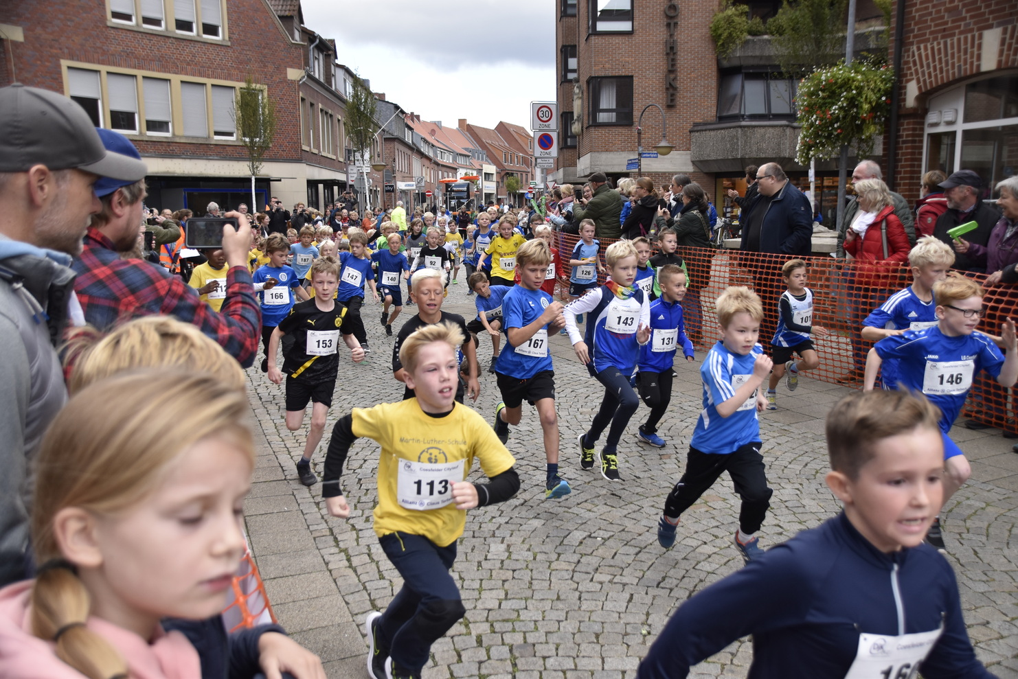 Eine Gruppe von Kindern in Sportkleidung läuft an einem Straßenschild entlang während eines Wettlaufs. Zuschauer beobachten.