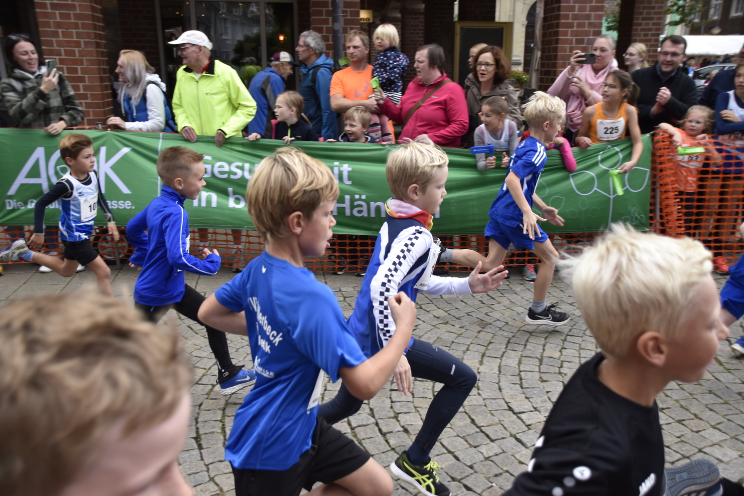 Gruppe von Kindern in blauen Trikots beim Laufen auf einer Straße, gekennzeichnet von Zuschauer*innen und einem Banner.