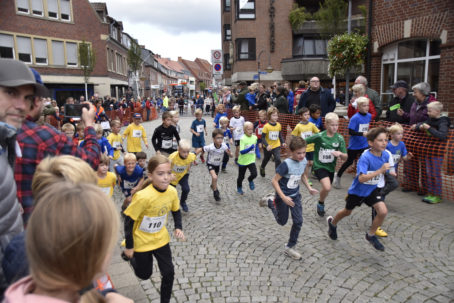 Gruppierung von Kindern in Sportbekleidung beim Start eines Straßenrennens, umgeben von Zuschauern in einer Stadt.