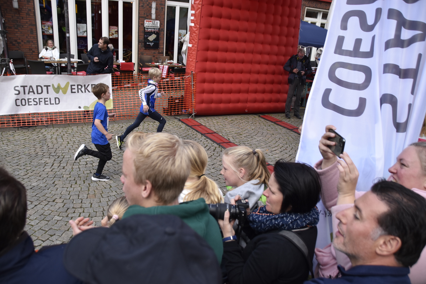 Zwei Kinderlaufen während eines Laufevents, umgeben von jubelnden Zuschauern und einer Banner.