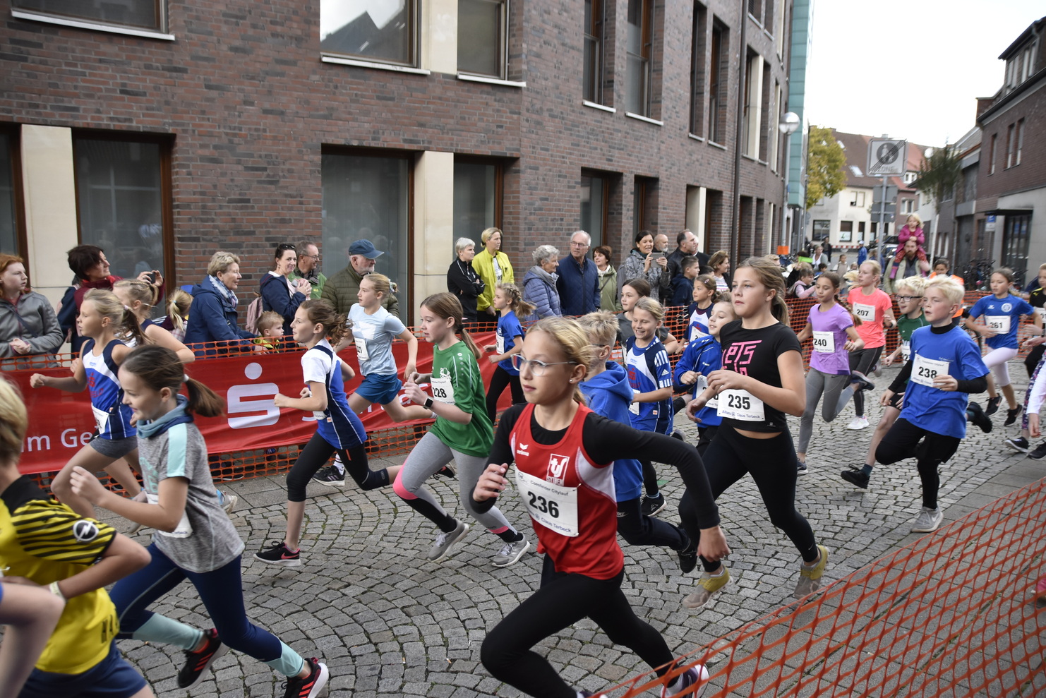 Gruppe von Mädchen in Sportkleidung beim Laufen in einer Stadt, Zuschauer stehen hinter einem Absperrgitter.