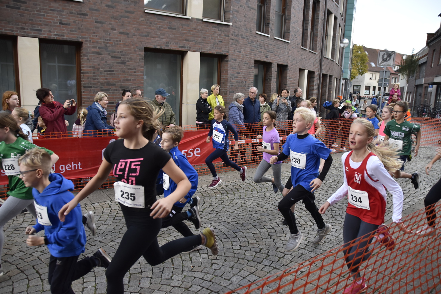 Gruppe von Kindern läuft im Wettkampf auf einer Steinpflasterstraße, umgeben von Zuschauern hinter einer Absperrung.