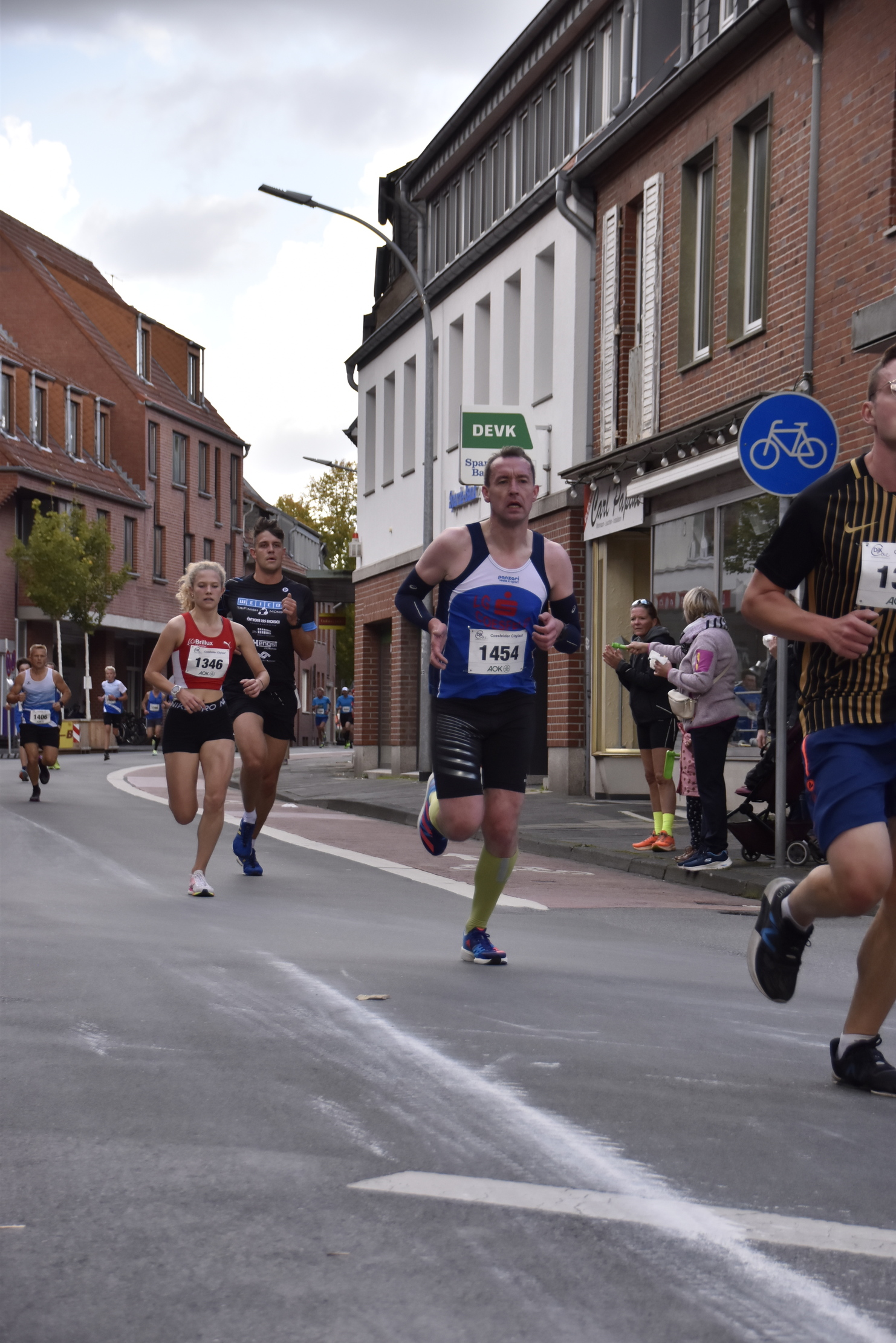 Vier Läufer in Wettkleidung laufen auf einer Straße, umgeben von Zuschauern und Gebäuden im Hintergrund.