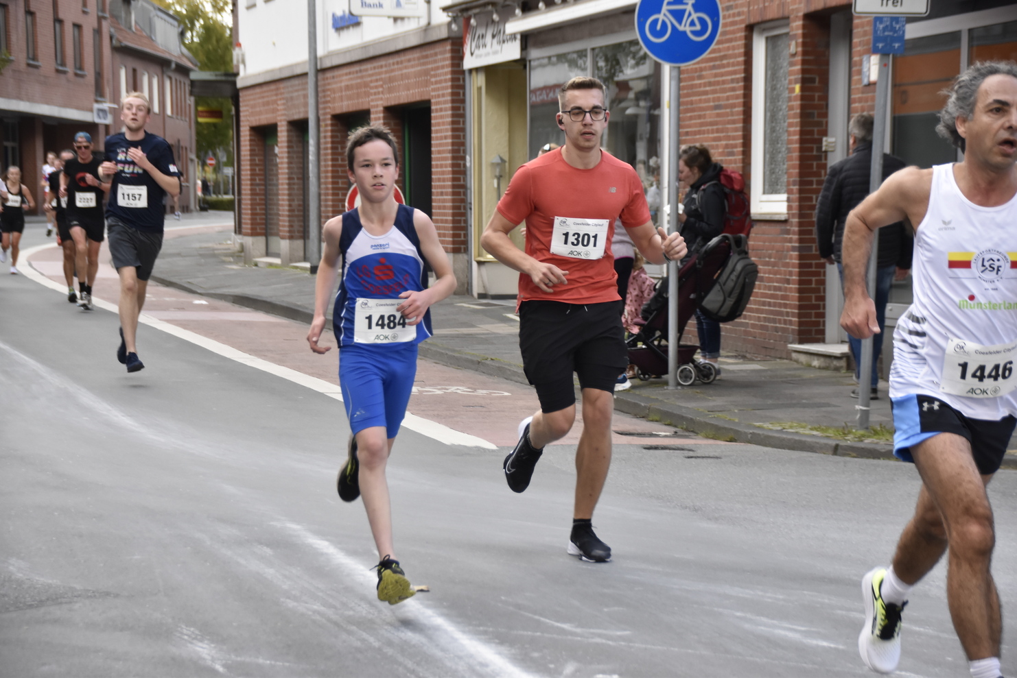 Verschiedene Läufer, darunter ein Junge in blauer Sportkleidung, laufen eine Straße entlang. Zuschauer stehen am Rand.