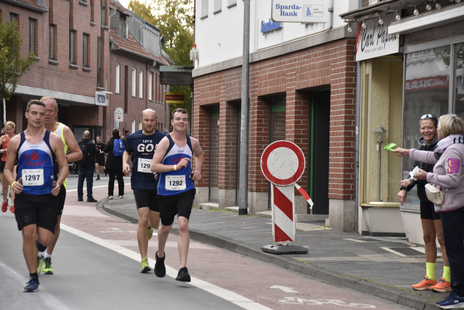 Vier Läufer in Sportkleidung joggen auf einer Stadtstraße, während Zuschauer ihnen zujubeln und Getränke reichen.