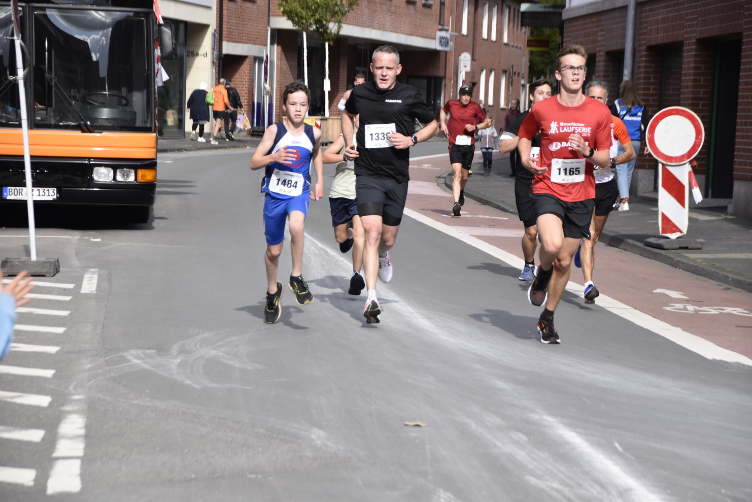 Vier Läufer in Sportkleidung nehmen an einem Straßenlauf teil; zuschauende Menschen und ein Bus im Hintergrund.