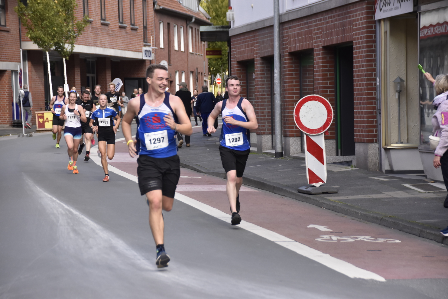 Läufer in Sportoutfits sprinten auf einer Straße, während Zuschauer am Rand anfeuern. Verkehrsschild im Hintergrund sichtbar.