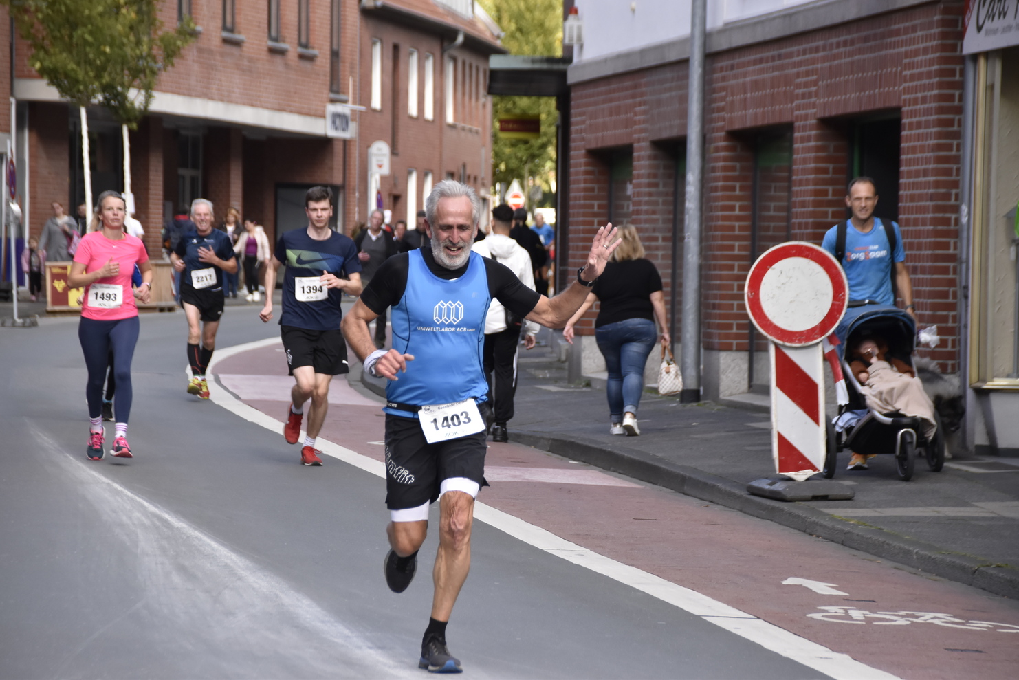 Läufer in sportlicher Kleidung winkt bei einem Wettbewerb auf einer städtischen Straße, umgeben von weiteren Teilnehmern.