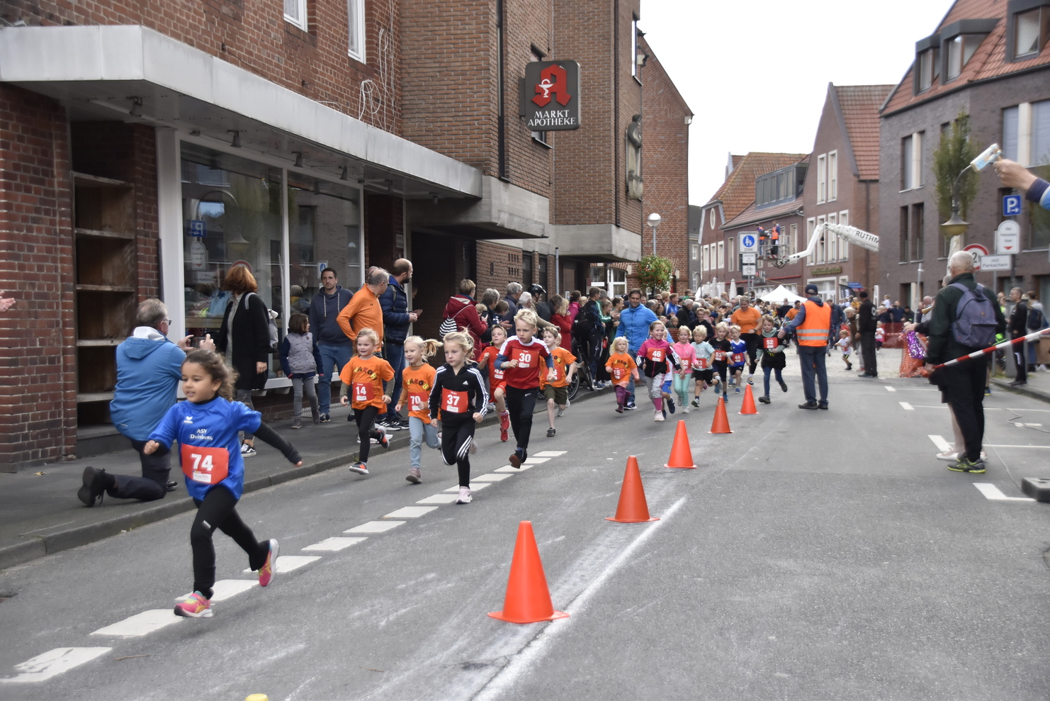 Kinder in bunten T-Shirts laufen einen Wettlauf auf einer Straße, umgeben von Zuschauern und orangefarbenen Pylonen.