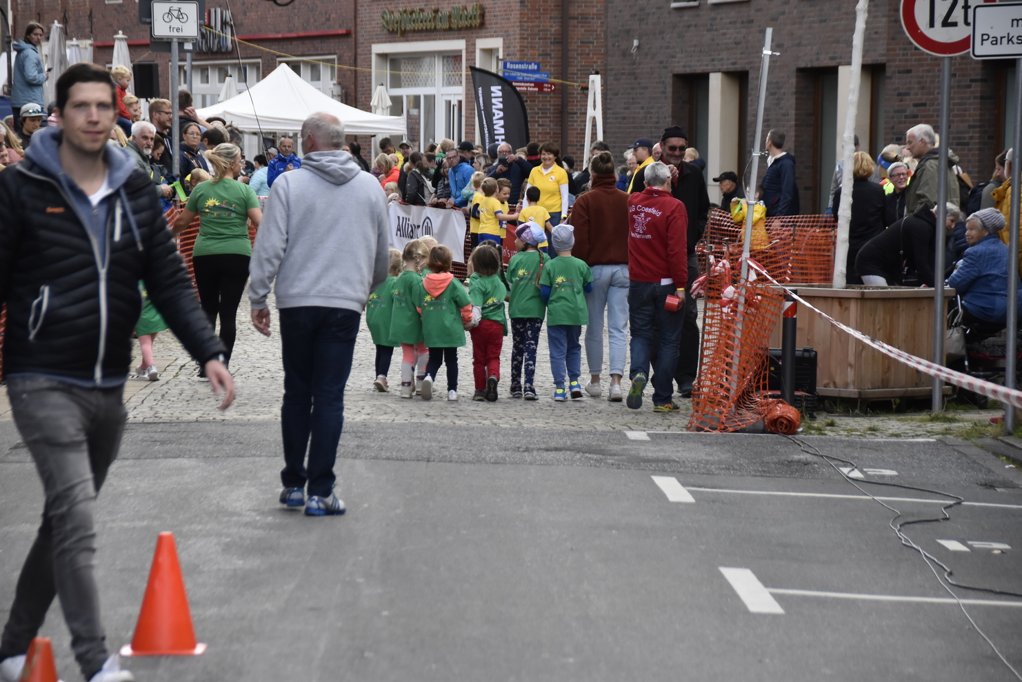 Gruppe von Kindern in grünen T-Shirts geht eine Straße entlang, begleitet von mehreren Zuschauern und Absperrungen.