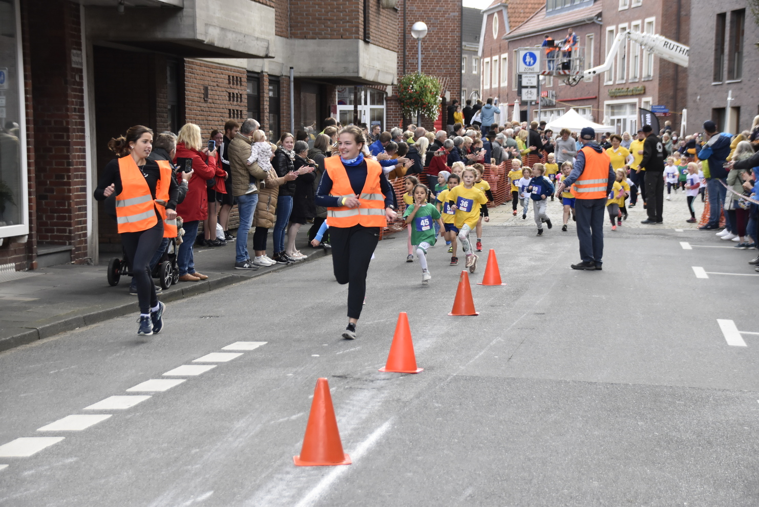 Kinderrennen auf einer Straße, mit Läufern in bunten Trikots und Zuschauern, die an der Strecke applaudieren.