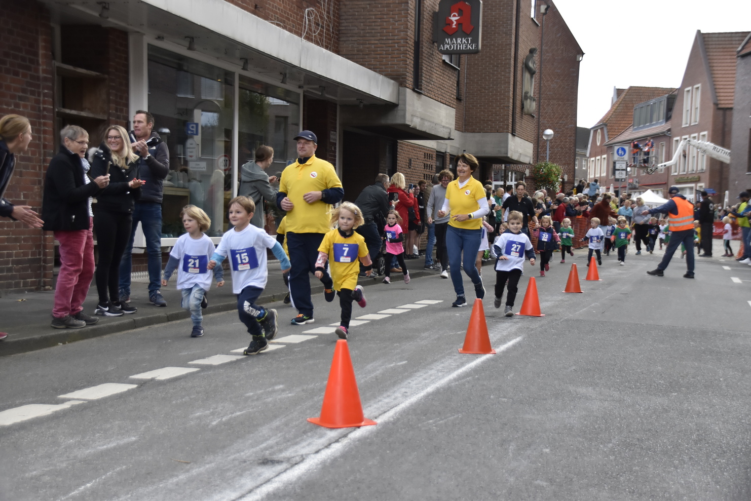 Kinder in Wettkampfkleidung laufen an Zuschauern vorbei, während eine Veranstaltung auf der Straße stattfindet.