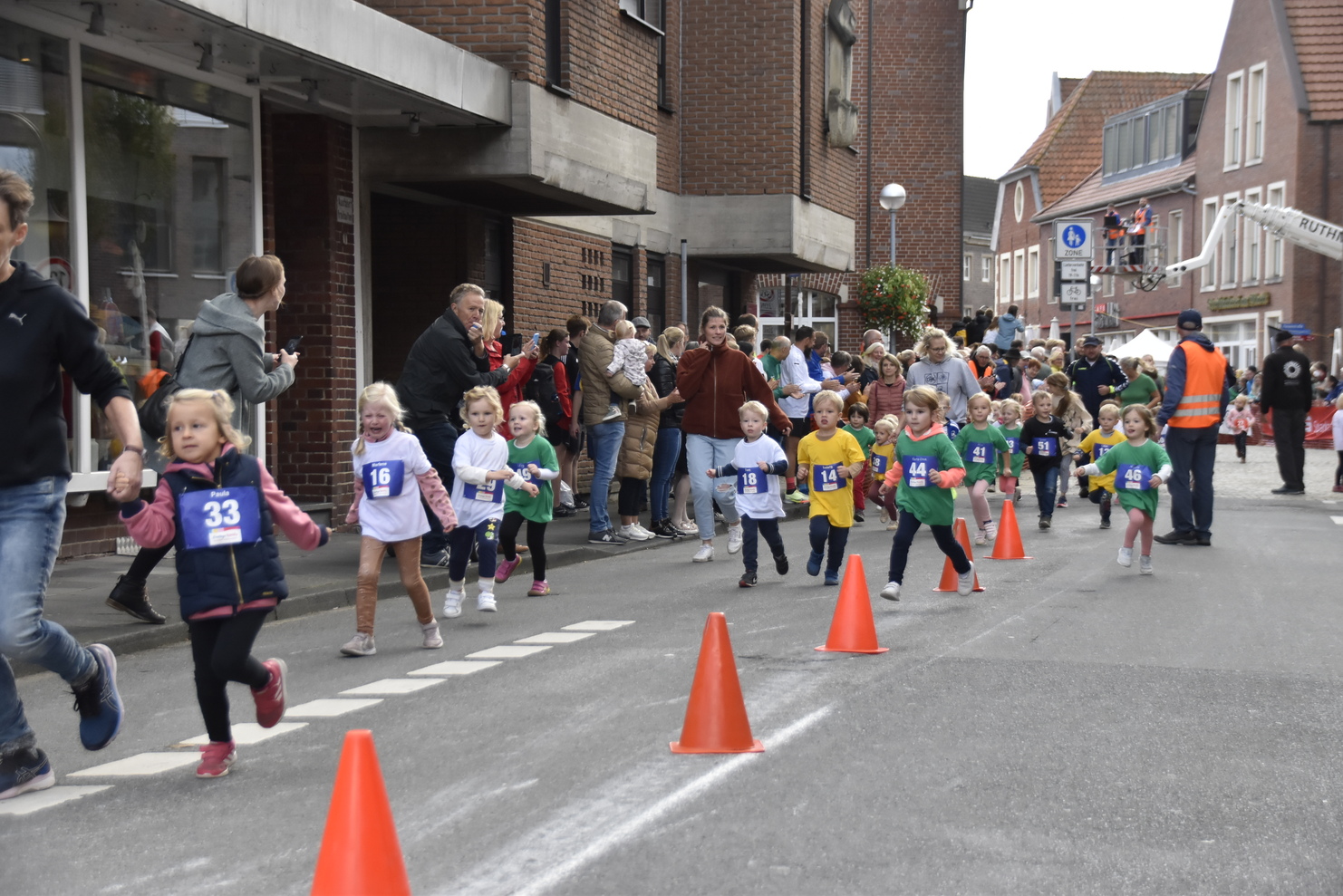 Gruppe von Kindern in Laufkleidung nimmt an einem Straßenlauf teil, umgeben von Zuschauern und Absperrungen.