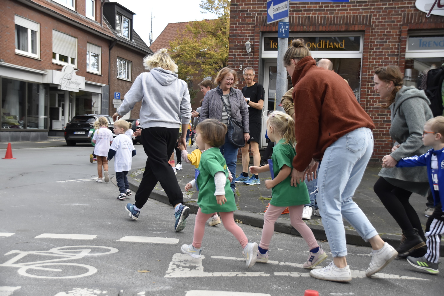 Gruppen von Kindern und Erwachsenen laufen gemeinsam auf einer Straßenkreuzung, umgeben von einheimischen Geschäften.