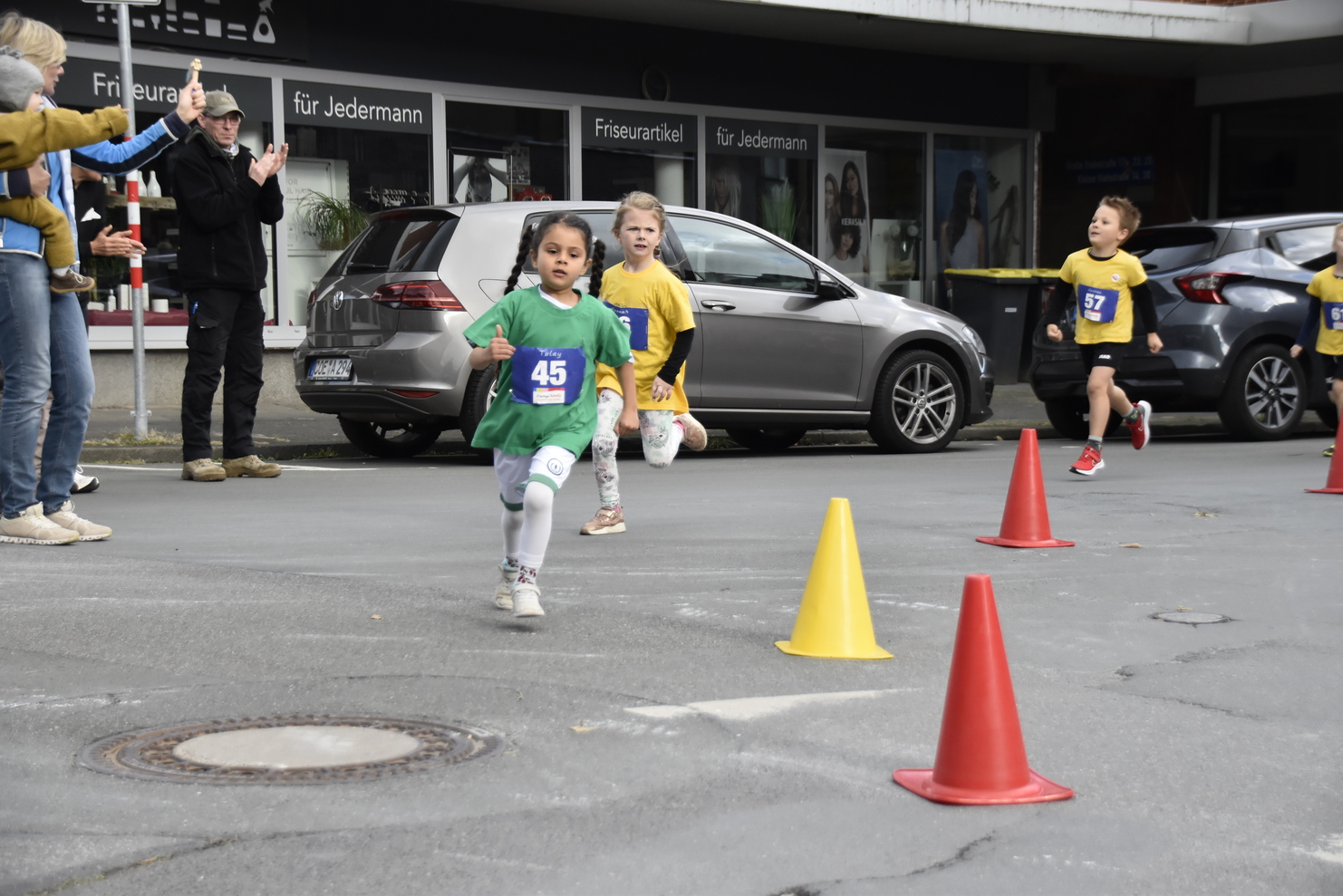 Kinder laufen beim Wettlauf durch einen markierten Streckenabschnitt mit Pylonen und jubelnden Zuschauern im Hintergrund.