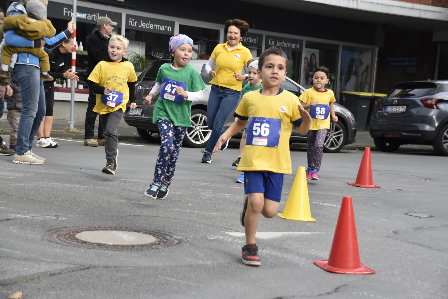 Kinder in Laufshirts nehmen an einem Wettlauf teil, während Zuschauer am Rand anfeuern. Mehrere Verkehrshütchen stehen bereit.