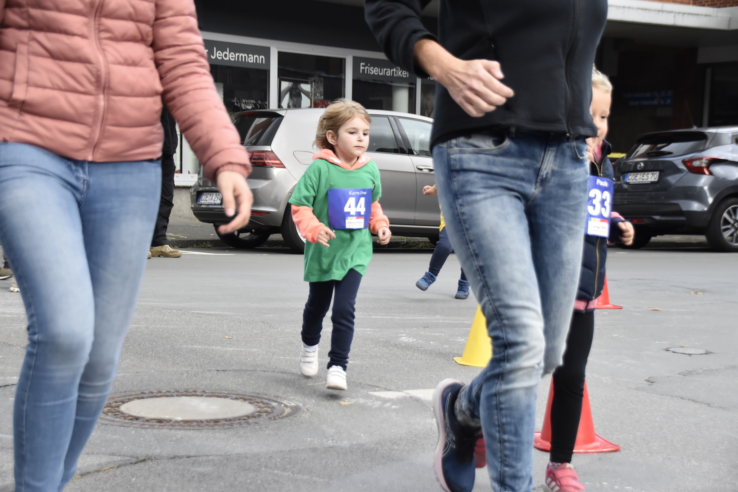 Kinder in Sportbekleidung laufen an einer Straße entlang, während Erwachsene im Hintergrund vorbeigehen.