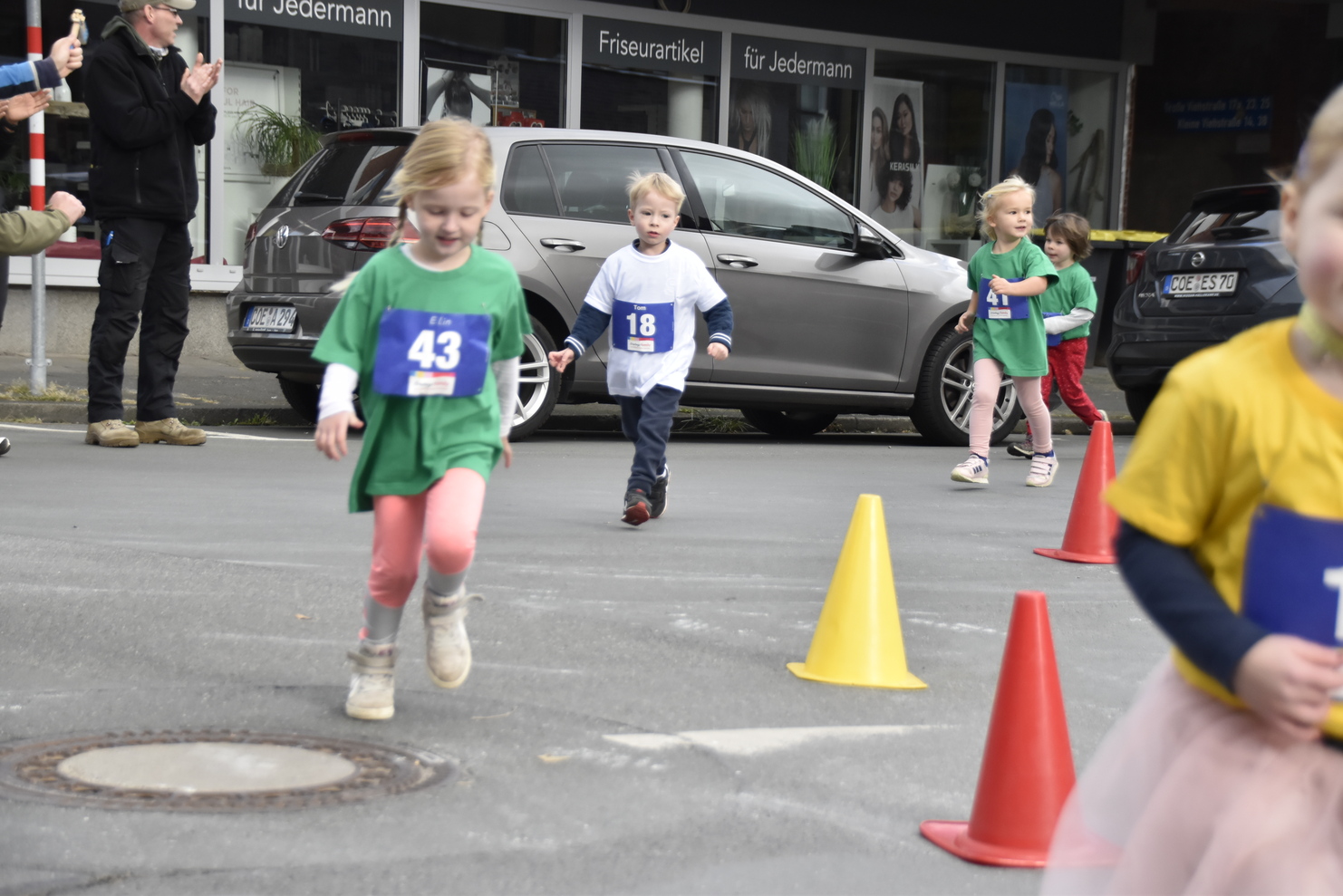 Kinder in Sportoberteilen laufen einen Wettlauf auf der Straße zwischen Pylonen, applaudierende Zuschauer im Hintergrund.