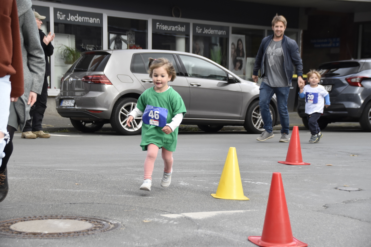 Ein Mädchen in grünem T-Shirt läuft fröhlich um orangefarbene Pylonen, während Erwachsene im Hintergrund stehen.