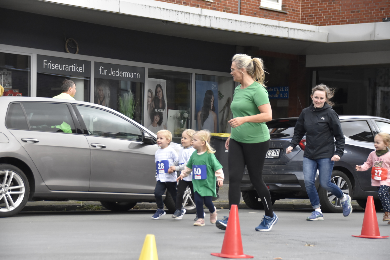 Kinder und Erwachsene laufen vor einem Friseursalon, gekennzeichnet durch bunte Startnummern und Verkehrscones.