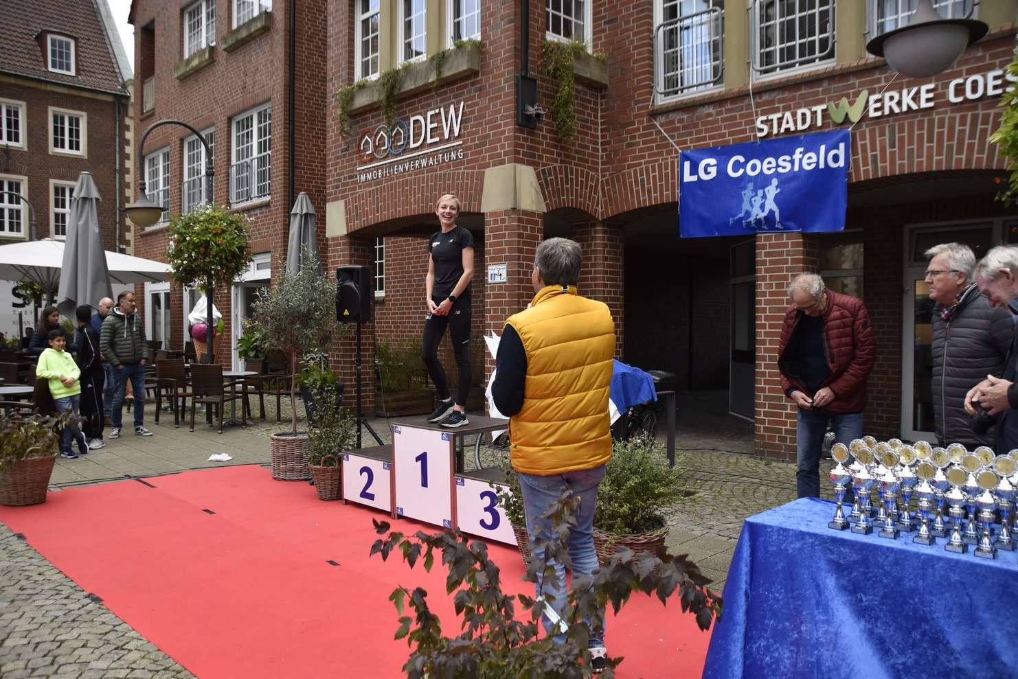 Frau auf dem Siegerpodest lächelt, während eine Gruppe Zuschauer und ein Redner ihr Anerkennung zollen.