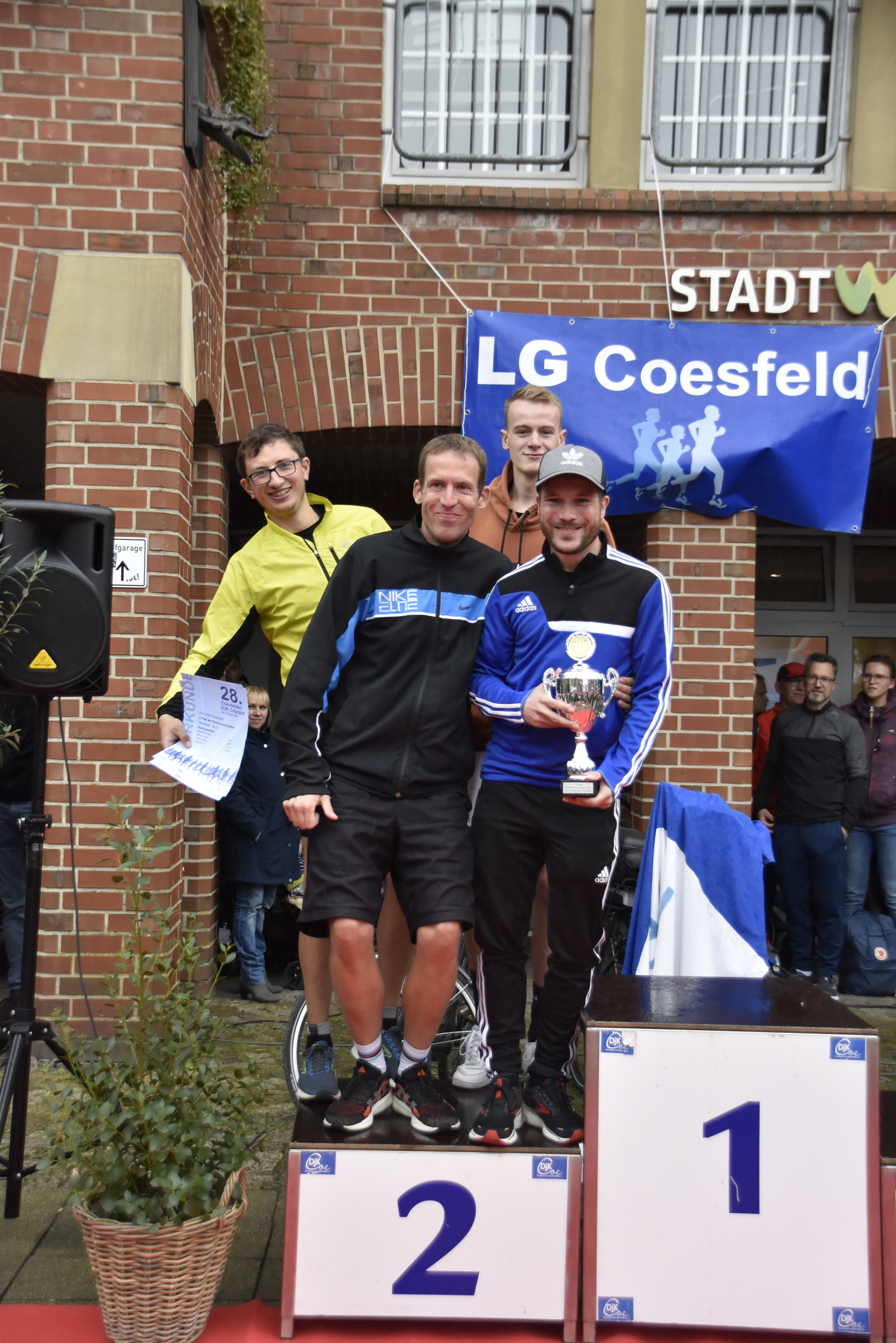 Fünf Männer feiern auf dem Siegertreppchen mit Pokal und Medaillen, im Hintergrund ein Banner der LG Coesfeld.