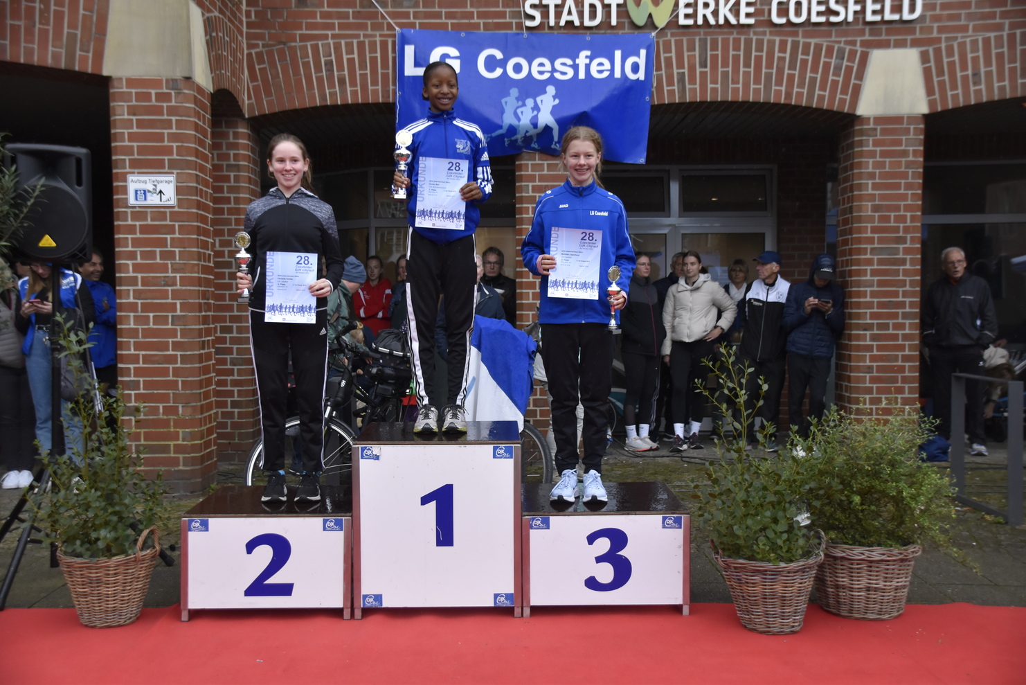 Drei Siegerinnen stehen auf dem Treppchen bei einer Sportveranstaltung, begleitet von Pokalen und Urkunden.