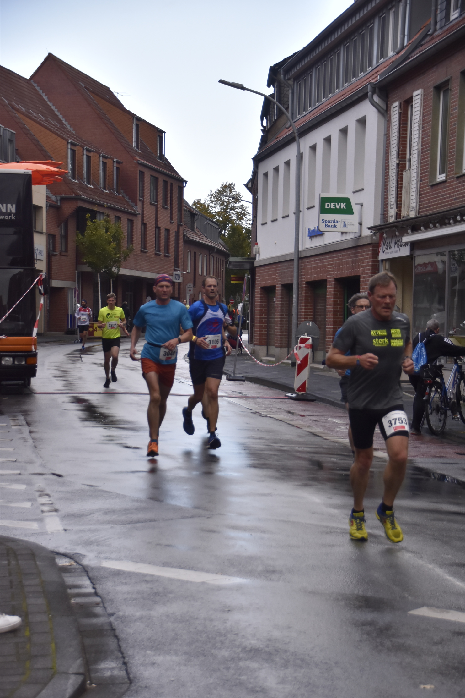 Teilnehmer eines Straßenlaufs laufen in einer nassen, schmalen Straße zwischen Häusern und verkehrsbedingt abgestellten Fahrzeugen.