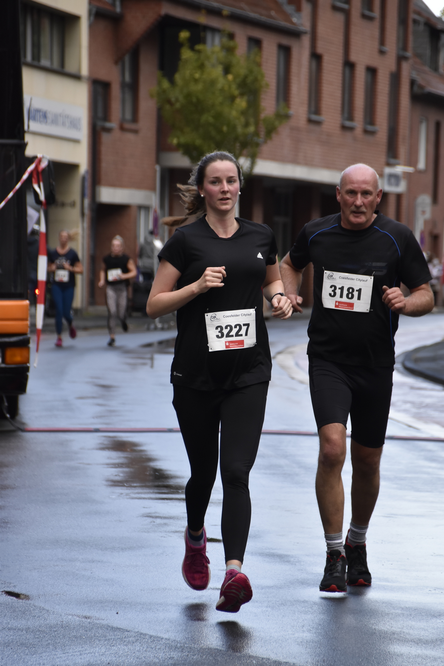 Zwei Läufer, eine Frau in Schwarz und ein Mann in Schwarz, joggen auf einer nassen Straße während eines Wettlaufs.