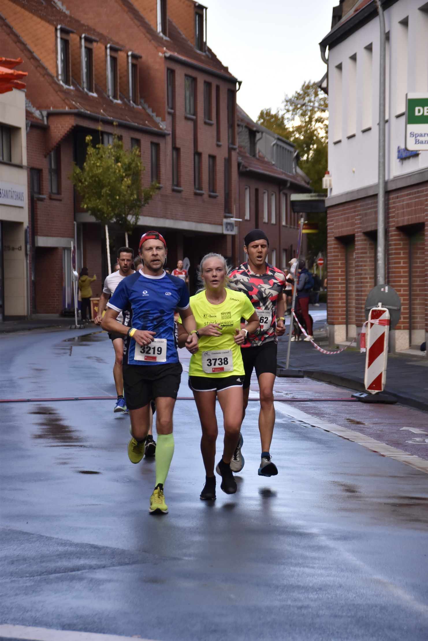 Vier Läufer in Sportbekleidung laufen auf einer nassen Straße zwischen Gebäuden während eines Wettkampfs.