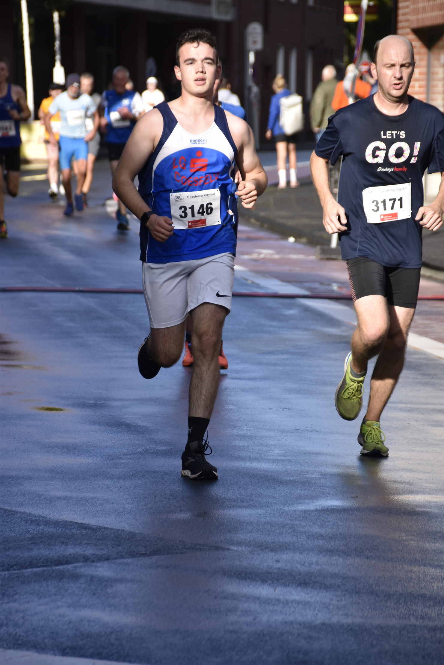 Zwei Läufer nehmen an einem Wettkampf teil, einer trägt ein blaues Trikot, der andere ein schwarzes T-Shirt mit Aufdruck.