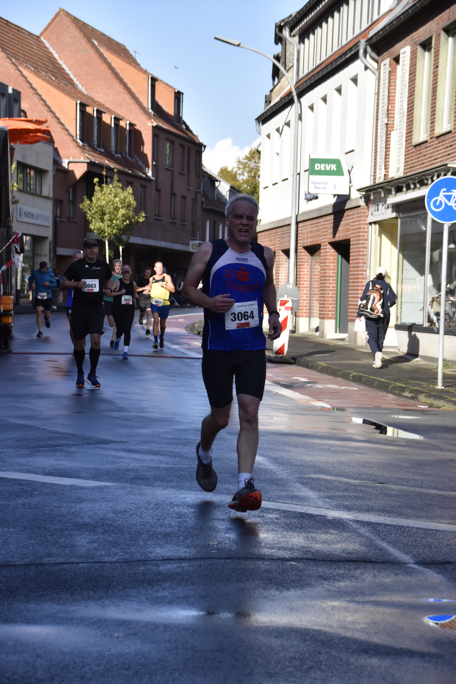 Läufer im blauen Shirt überquert eine nasse Straße während eines Wettkampfs, weitere Teilnehmer im Hintergrund.