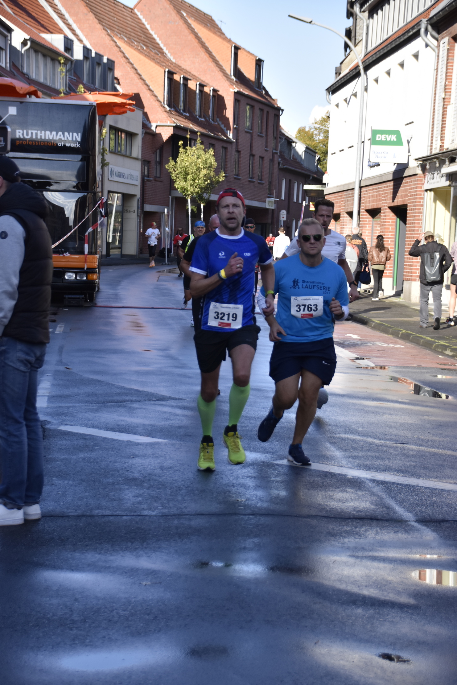 Zwei Läufer in Sportbekleidung joggen auf einer nassen Straße, vorbei an Zuschauern und einem Lkw.