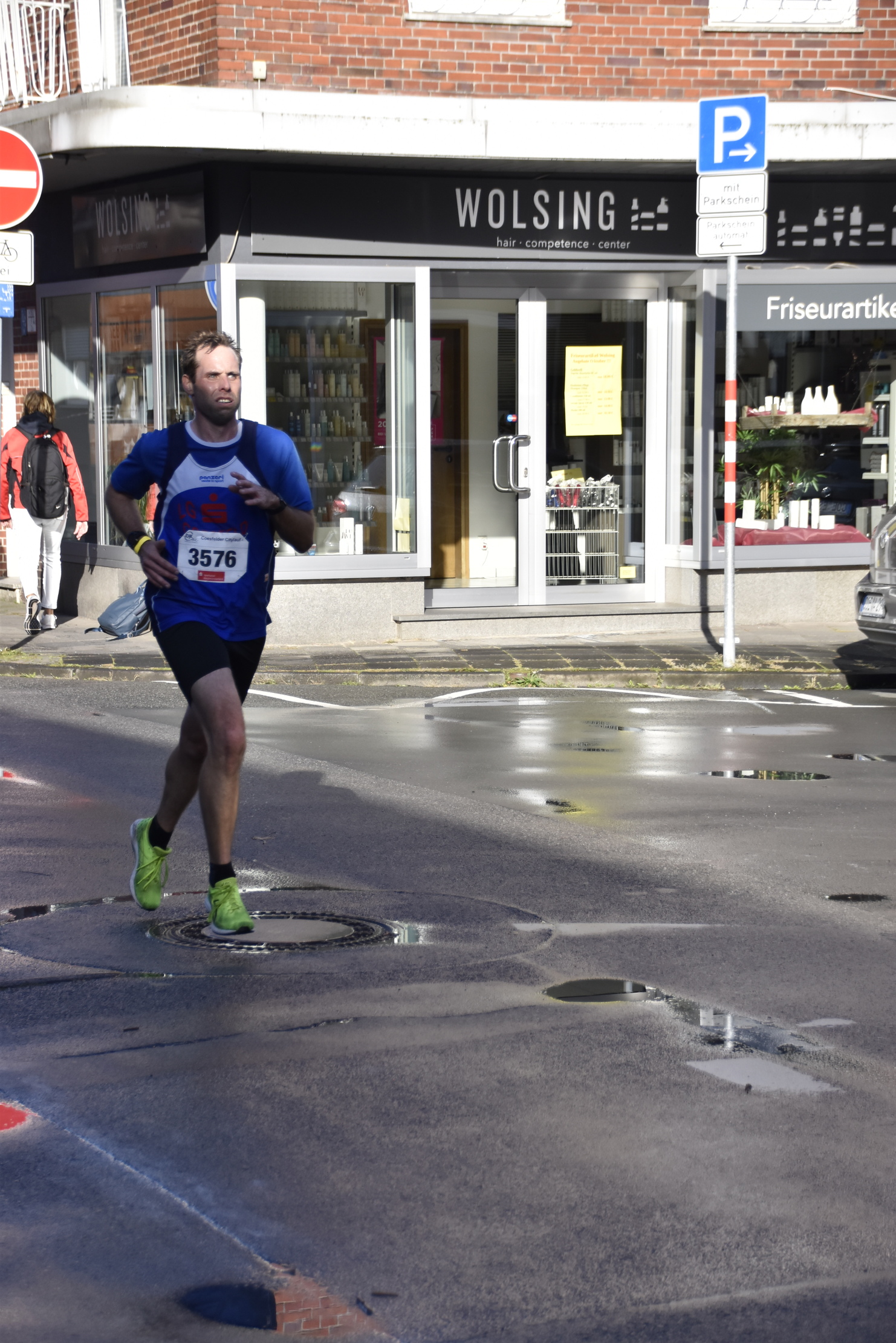 Läufer in blauem Shirt mit Startnummer 3576 joggt auf nasser Straße vor einem Friseursalon.