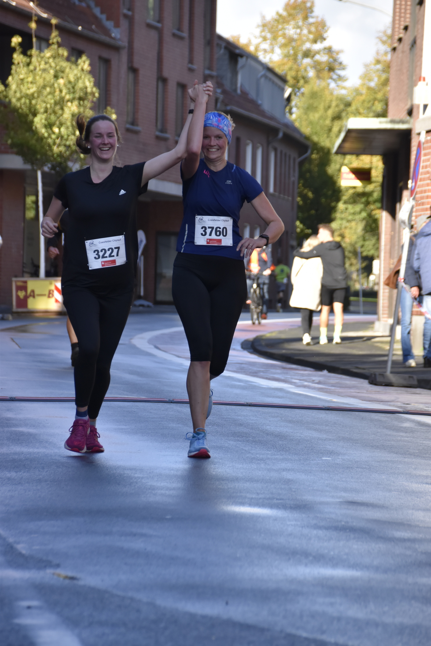Zwei Frauen jubilieren beim Überqueren der Ziellinie eines Straßenlaufs, Hände erhoben und lächelnd.